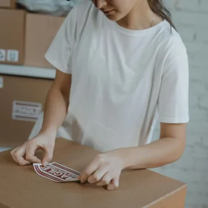 Woman preparing shipment by applying a 'fragile' sticker on a cardboard box in a warehouse.