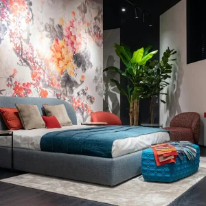 Comfortable bed with colorful cushions and blue blanket placed at painted wall in modern bedroom with armchairs and potted plant